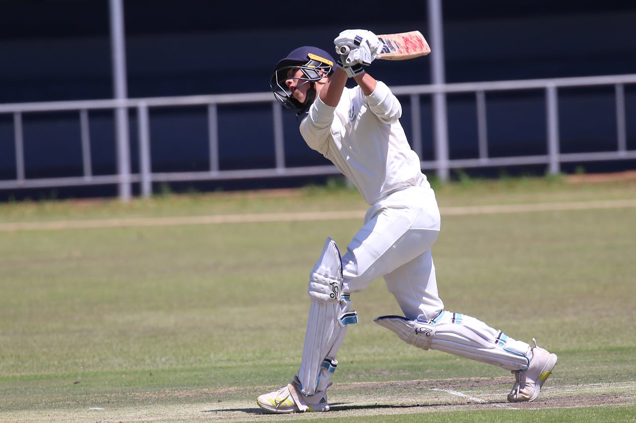Cricketer in action hitting a shot on an outdoor field during a sunny day.